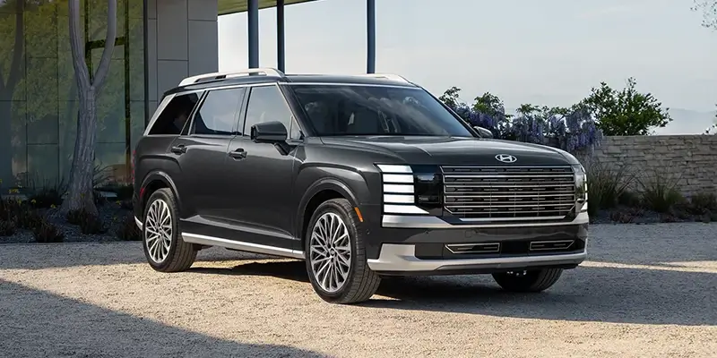 Dark gray 2026 Hyundai Palisade Hybrid with a wide chrome grille, stacked LED headlights, large alloy wheels, and a boxy design, parked on a gravel driveway beside a modern glass building with trees in the background.