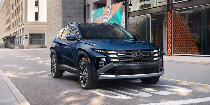 Blue 2026 Hyundai Tucson driving on a city street past modern buildings, featuring a parametric grille with integrated LED lights, black trim, and alloy wheels.