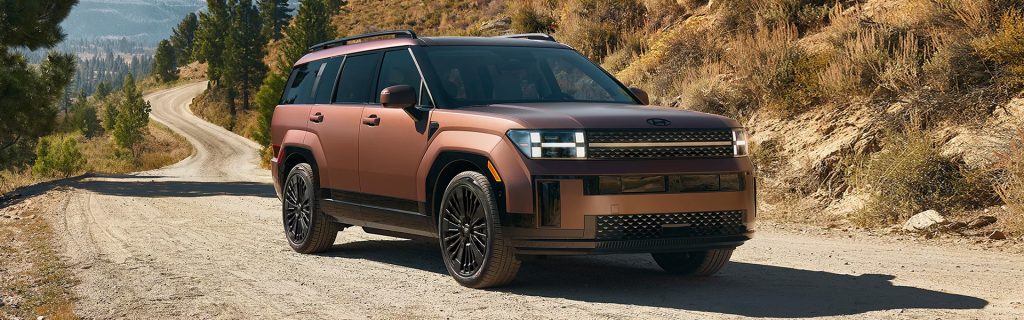 Matte brown 2026 Hyundai Santa Fe driving on a winding dirt road through a mountainous landscape with trees and dry hills, featuring a bold front grille and modern LED headlights.