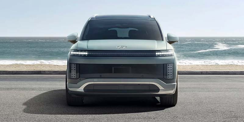 A front-facing view of a modern Hyundai vehicle parked along a coastal road, featuring a bold, boxy design with a wide LED light bar, squared-off grille, and the ocean stretching out in the background.
