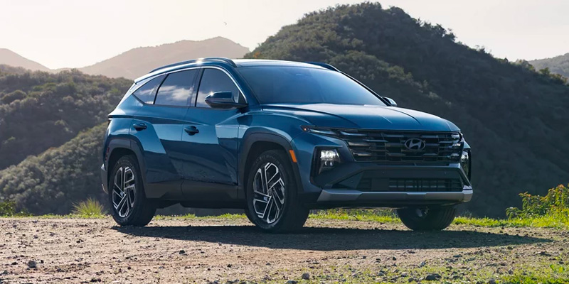 A blue Hyundai Tucson parked on a rugged dirt overlook, with rolling green hills and mountains in the background under soft daylight.