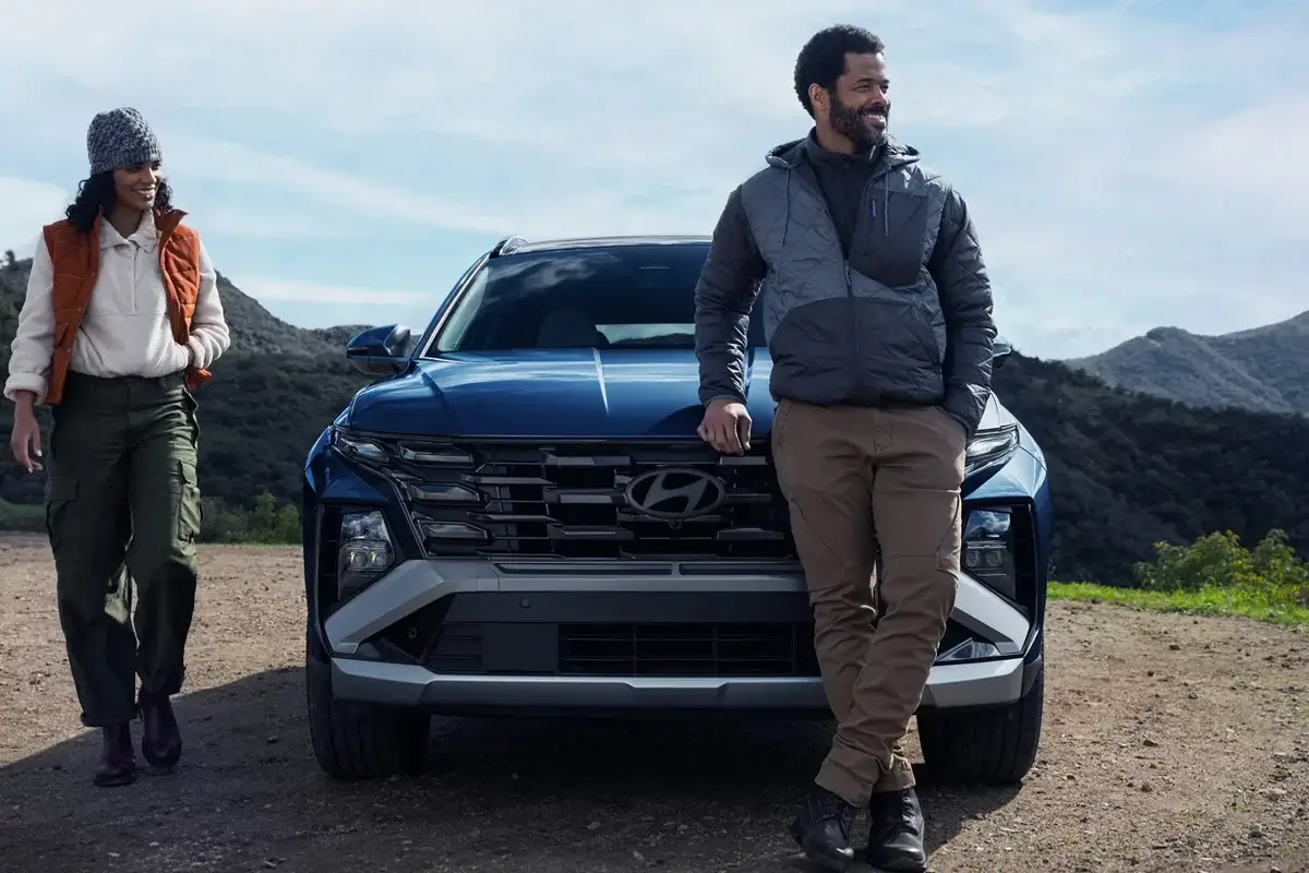 A man and a woman stand near a blue Hyundai vehicle in a scenic outdoor setting with mountains in the background.