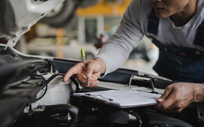 A mechanic working on a car engine while taking notes on a clipboard.