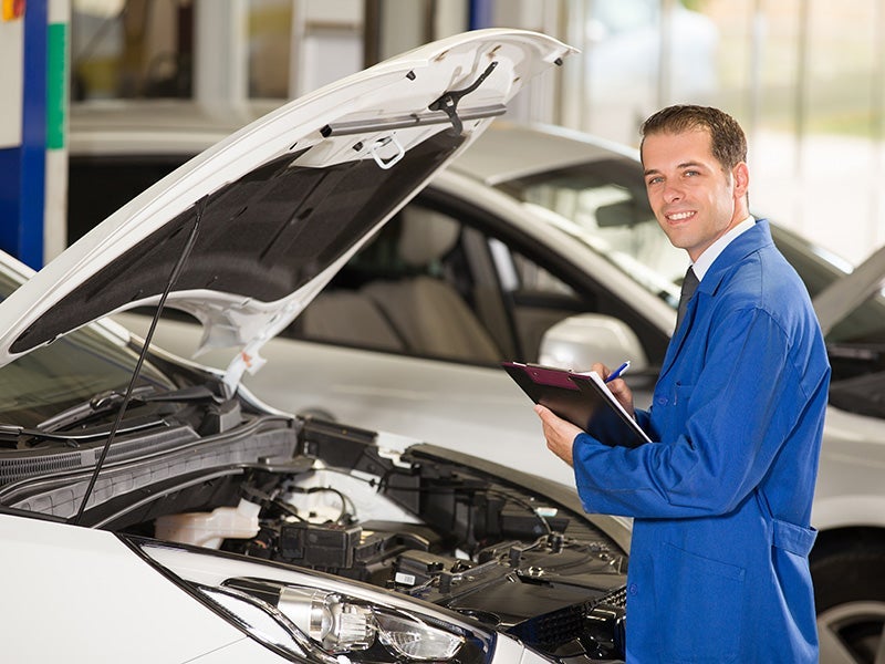 A mechanic in a blue uniform standing next to a car with the hood open, holding a clipboard and smiling.
